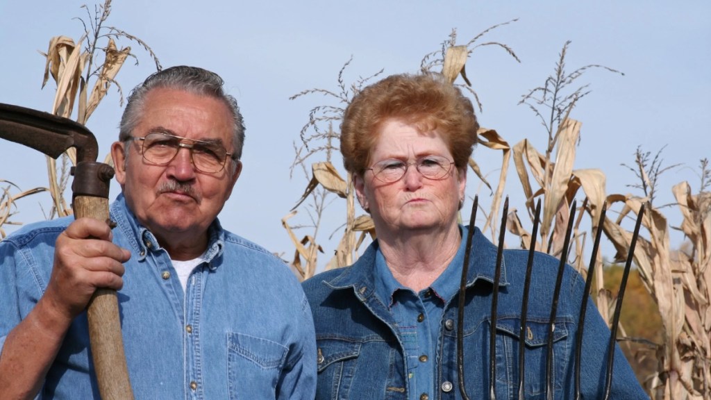 Ein Bauer steht mit seiner Frau mit grimmigem Gesichtsausdruck vor einem Maisfeld.