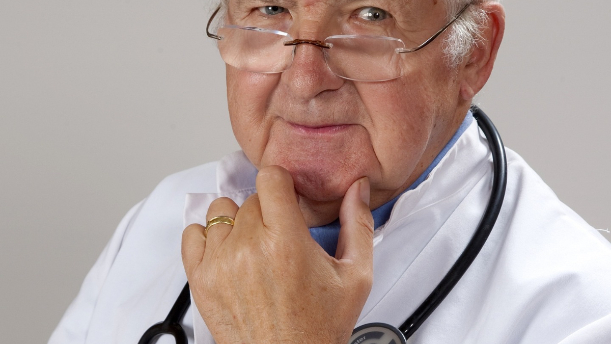 Ein älterer Arzt mit Stethoskop um den Hals und weißem Kittel schaut kritisch in die Kamera.