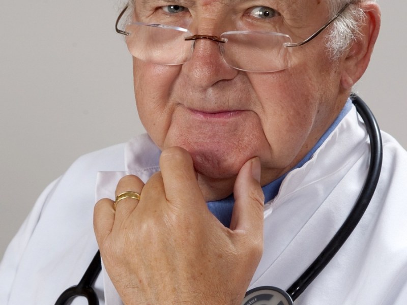 Ein älterer Arzt mit Stethoskop um den Hals und weißem Kittel schaut kritisch in die Kamera.