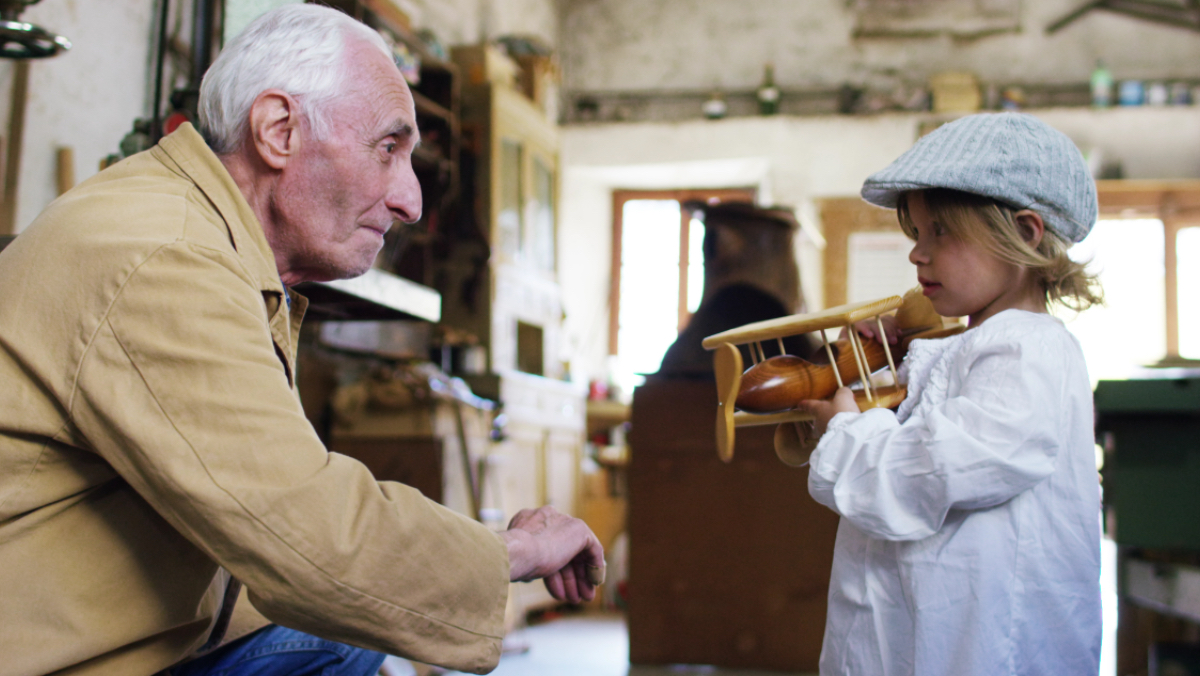 Ein Rentner in einer Werkstatt gibt einem kleinen Jungen ein Holzflugzeug.