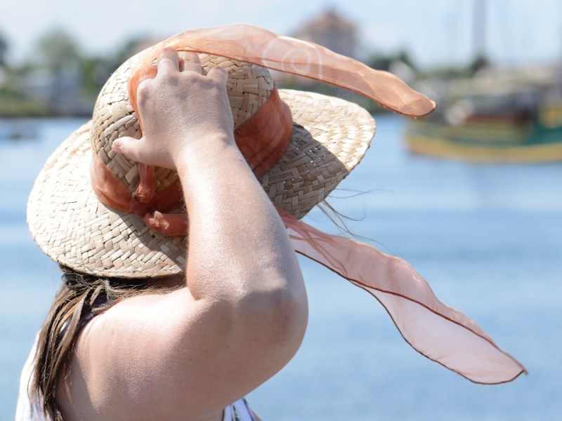 Eine ältere Dame steht an einem Hafen und hält ihren Hut fest.