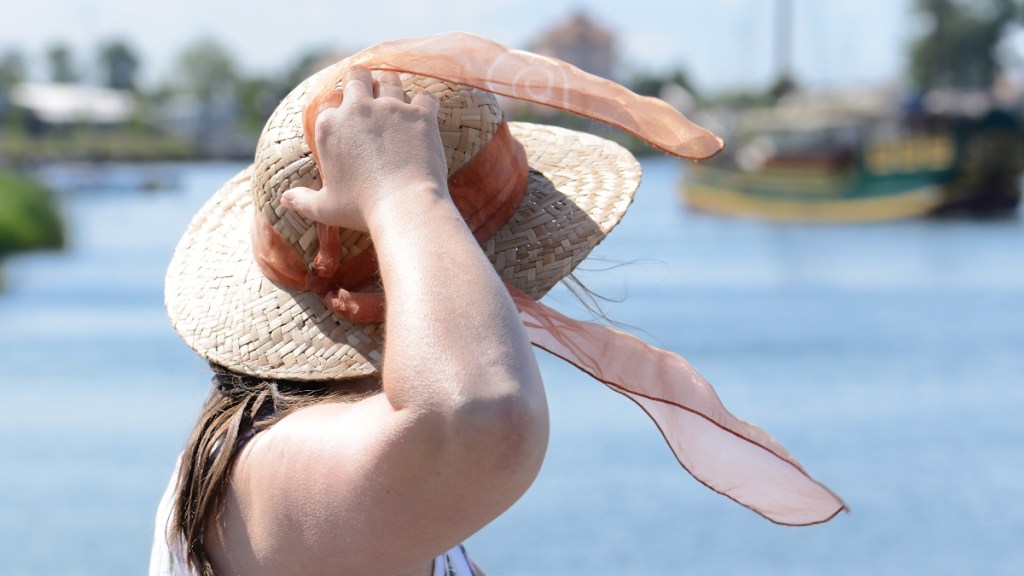 Eine ältere Dame steht an einem Hafen und hält ihren Hut fest.