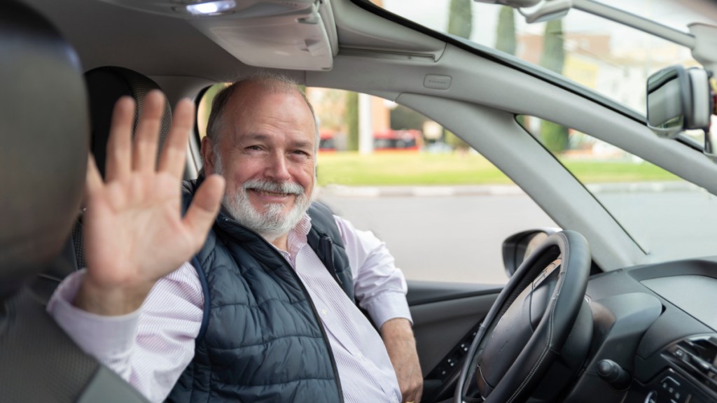 Ein Foto von einem älteren Taxifahrer, der hinter seinem Steuer sitzt und in die Kamera winkt.