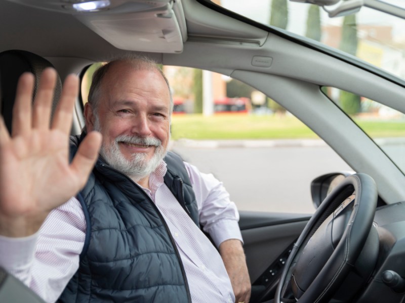 Ein Foto von einem älteren Taxifahrer, der hinter seinem Steuer sitzt und in die Kamera winkt.
