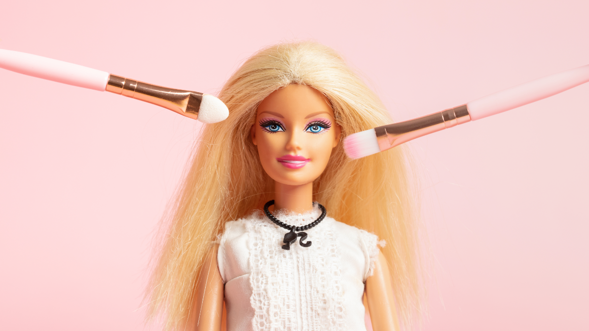Eine Barbie-Puppe mit Schminkpinseln vor dem Gesicht.
