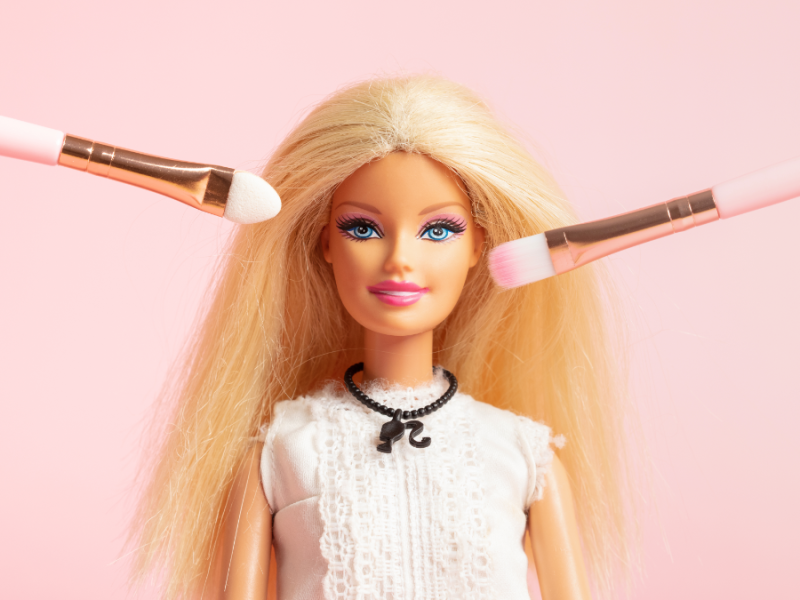 Eine Barbie-Puppe mit Schminkpinseln vor dem Gesicht.