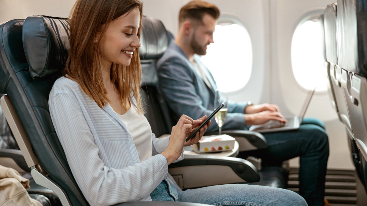 Fröhliche weibliche Person, die eine SMS auf ihrem Smartphone schreibt und lächelt, während sie im Passagiersessel im Flugzeug sitzt.