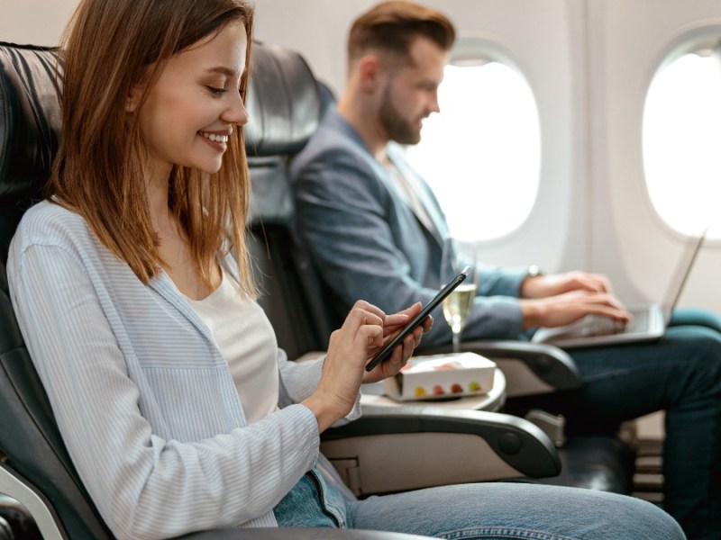 Fröhliche weibliche Person, die eine SMS auf ihrem Smartphone schreibt und lächelt, während sie im Passagiersessel im Flugzeug sitzt.